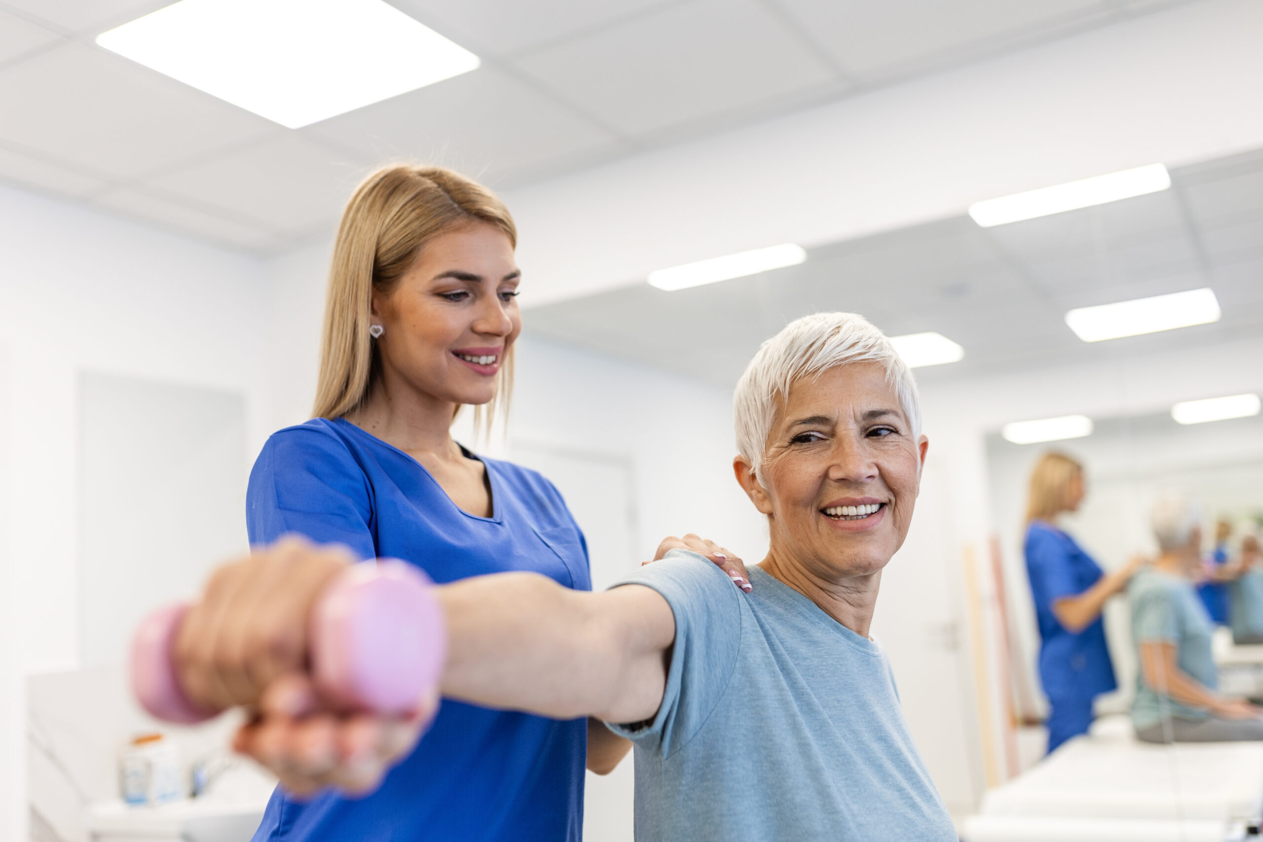 A therapist helps a smiling older woman lift a pink dumbbell during exercise.