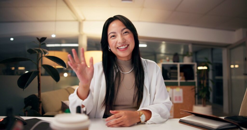 A young woman smiles and waves at the camera during a video call.