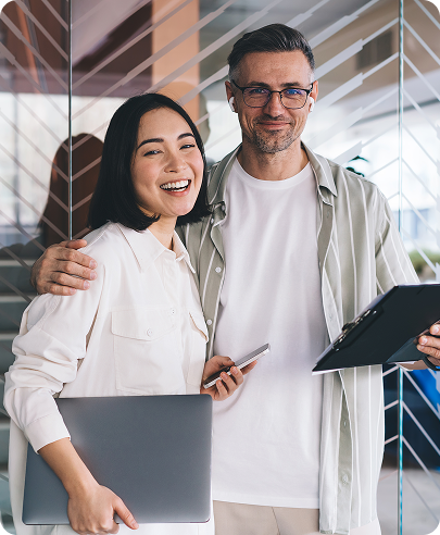 A smiling man and woman stand together holding a laptop and clipboard in a modern office.