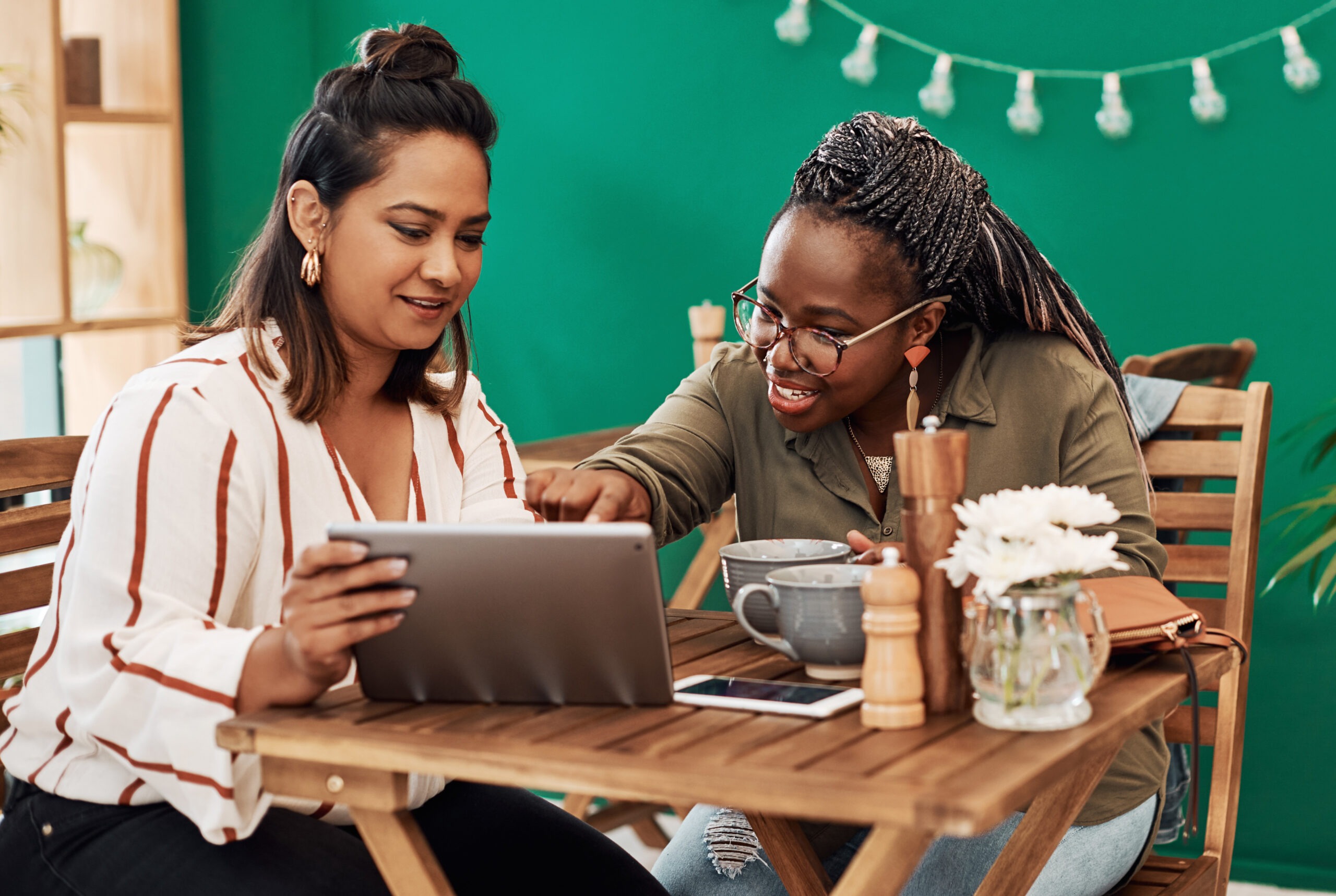 Two smiling women sit at a wooden table and look at a tablet.