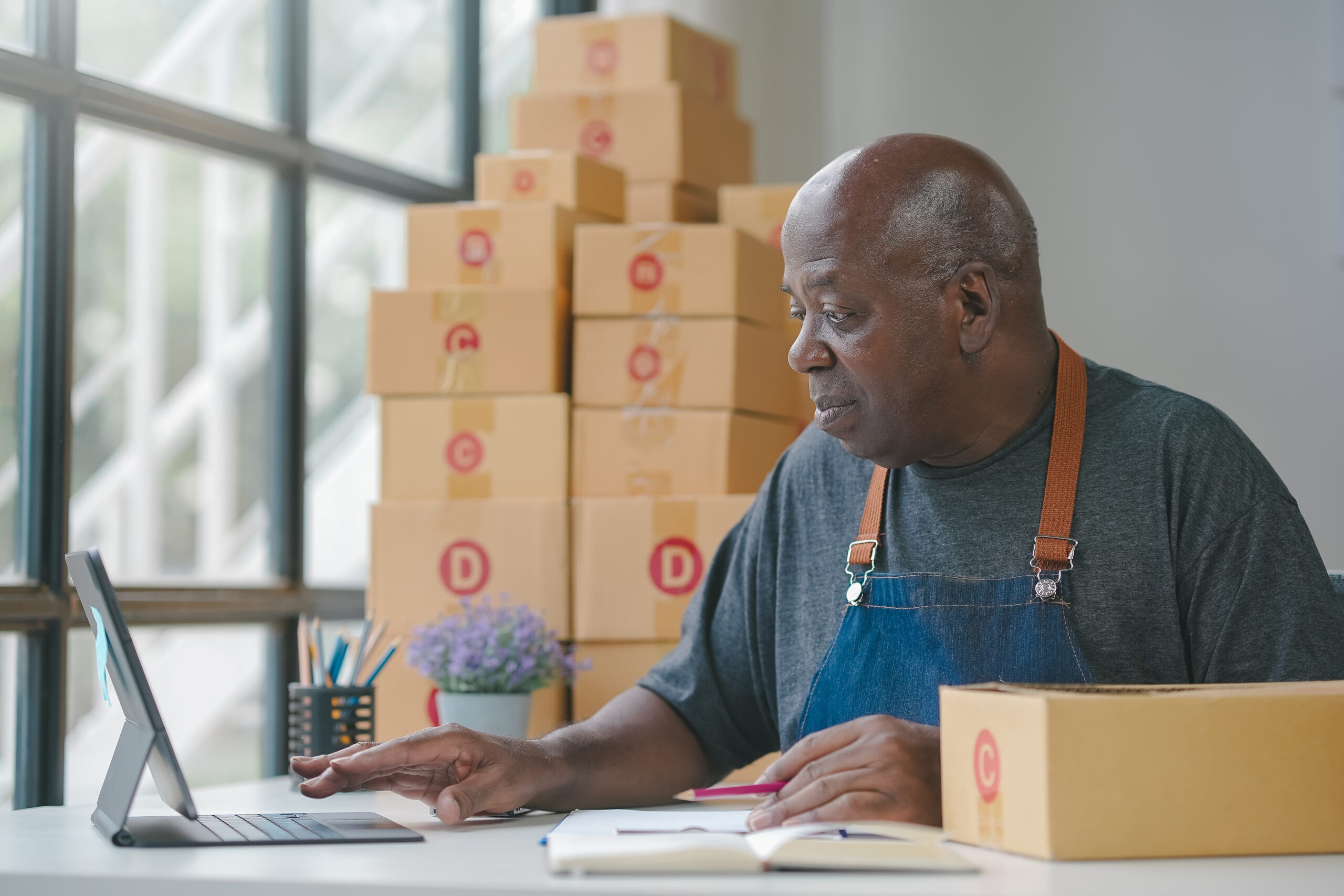 Senior man in workshop working on a laptop with boxes stacked in the background, focused and diligent in his task.