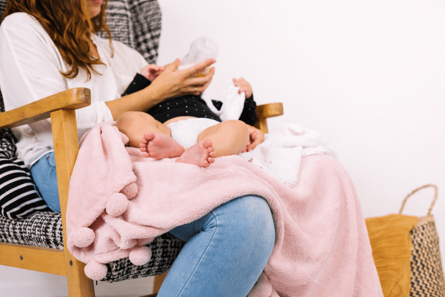 Adult seated in a chair bottle‑feeding a baby wrapped in a pink blanket.