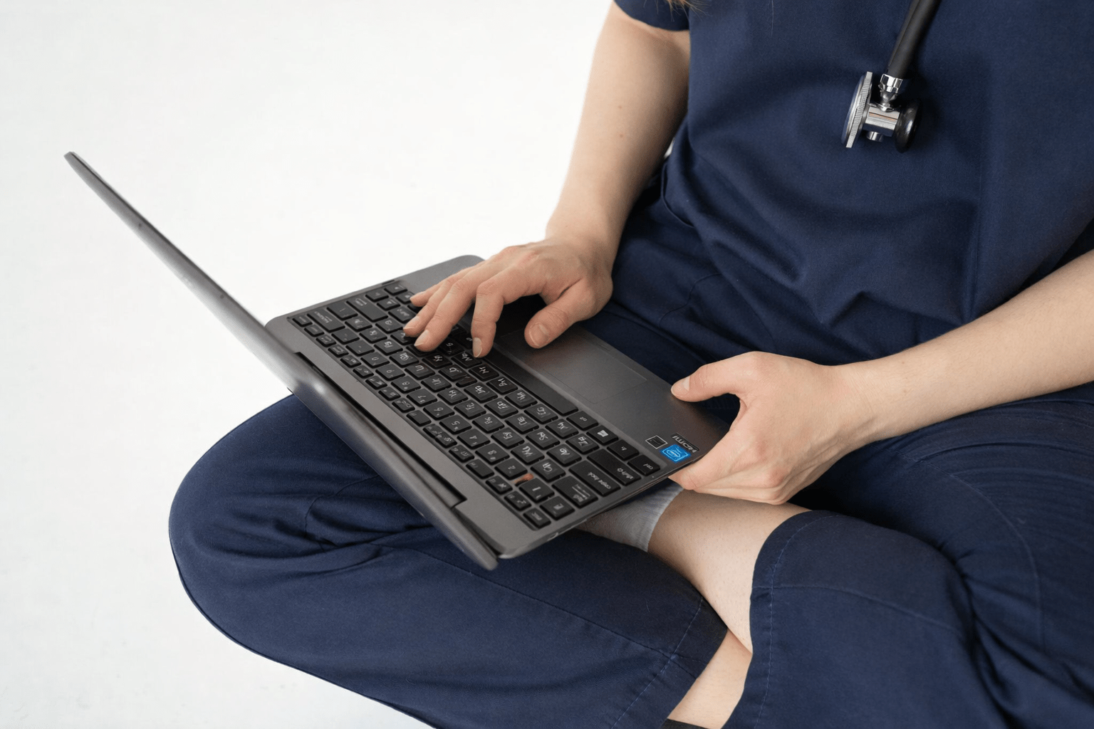 Medical professional in scrubs sitting cross‑legged while working on a laptop.